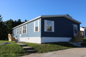 A blue single-wide manufactured home with white skirting and wooden steps under a clear sky.