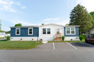 A one-story, blue and white manufactured home with a front porch and steps, situated in a residential community with trees and a car parked nearby.
