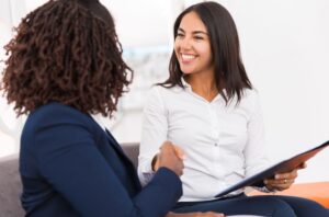 Two women in business attire sit and shake hands in an office setting. One holds a folder and smiles warmly, reflecting the professionalism and connection fostered at Aspire Communities.