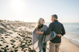 An older couple walks arm in arm along a sandy beach near the shoreline on a sunny day, enjoying the peaceful atmosphere that Aspire Communities are known for.