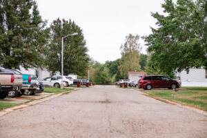 A paved Riverwood residential street lined with parked cars, green lawns, trees, and white houses on both sides under an overcast sky.