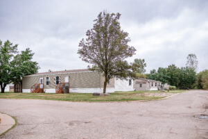 A row of manufactured homes sits along a quiet Riverwood street, with trees and cloudy skies overhead.