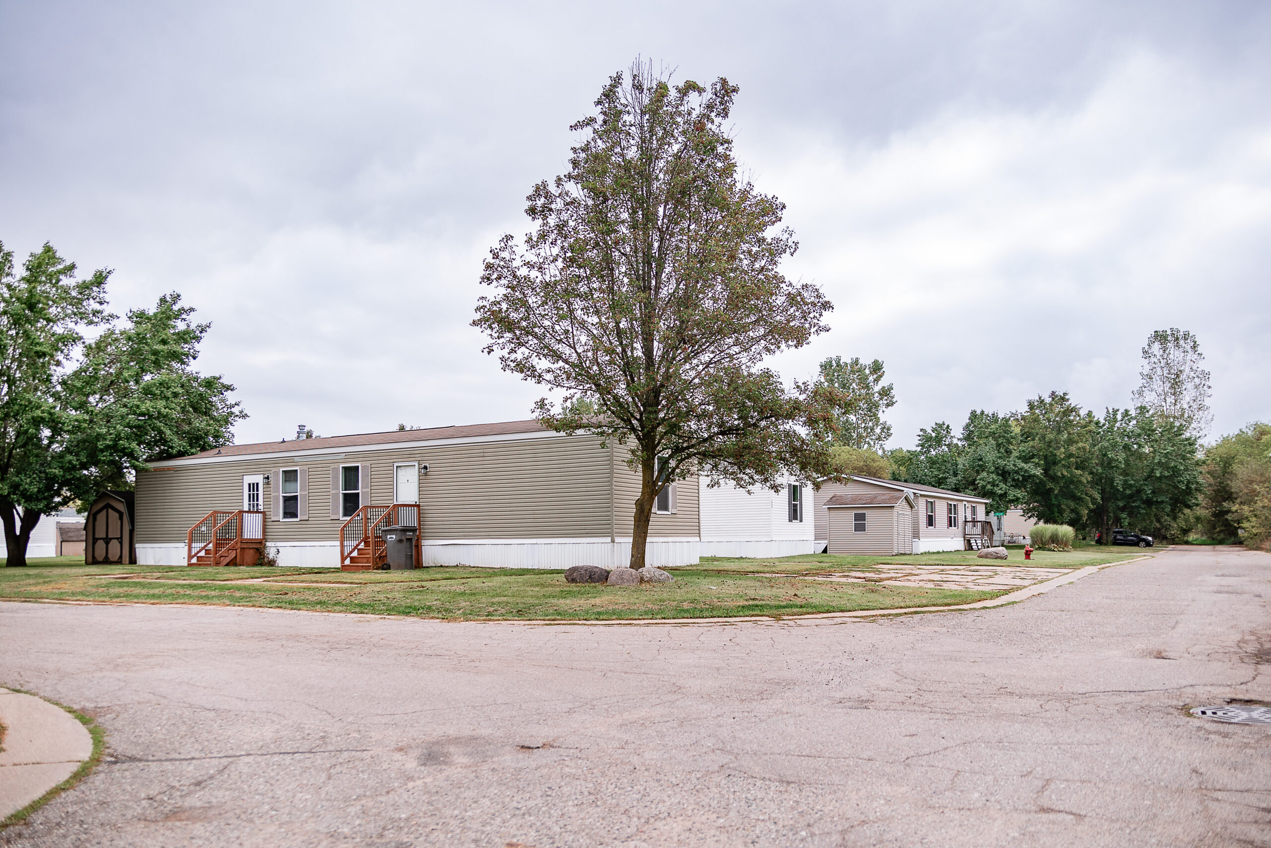 A row of manufactured homes sits along a quiet Riverwood street, with trees and cloudy skies overhead.