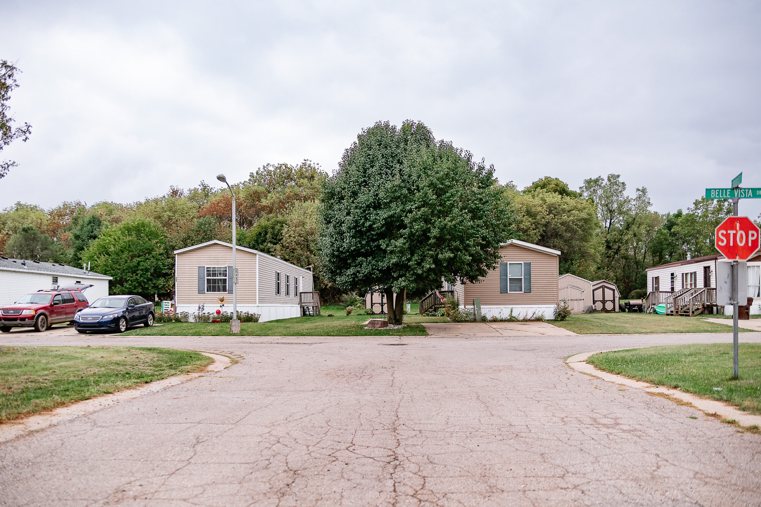 A suburban street corner in Riverwood, with a stop sign, two manufactured homes, a large tree at the center, and parked vehicles beneath an overcast sky.