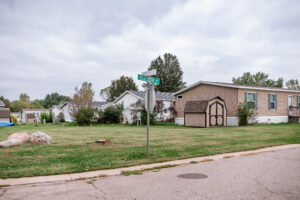 A street sign at a corner in the Riverwood neighborhood with manufactured homes, sheds, grass lawns, and trees under a cloudy sky.