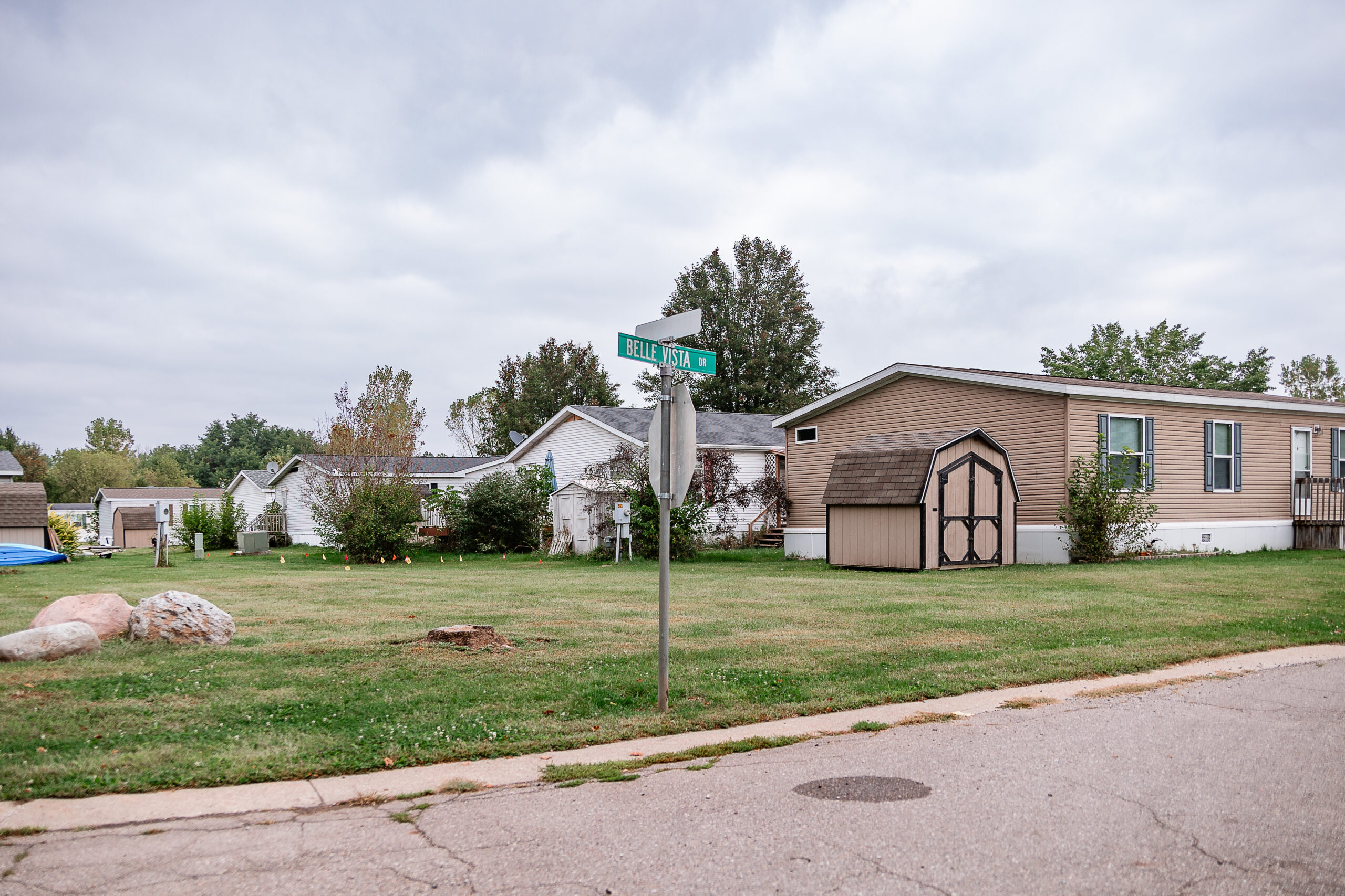 A street sign at a corner in the Riverwood neighborhood with manufactured homes, sheds, grass lawns, and trees under a cloudy sky.
