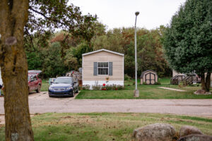 A beige Riverwood mobile home with shutters, a garden in front, a car in the driveway, a backyard shed, and trees surrounding the area.