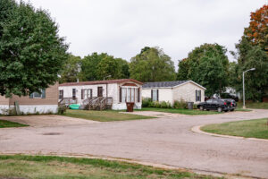 A residential street in Riverwood with mobile homes, parked cars, leafy trees, and a cloudy sky.