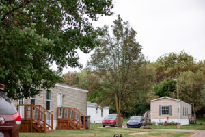 Two Riverwood mobile homes with wooden steps are surrounded by trees and parked cars on a grassy lot under an overcast sky.