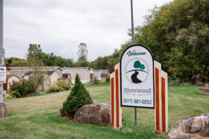 A sign reading "Welcome Riverwood (517) 881-0253" stands on a grassy area in Riverwood, with homes and trees in the background.