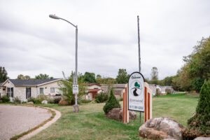 A street corner with a “Welcome to Riverwood” sign, a 15 mph speed limit sign, and a lamp post stands beneath a cloudy sky, with several Riverwood houses visible in the background.