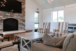 A living room in Lakeview Village features striped armchairs, a beige sofa, a light wood coffee table, a stone fireplace, and large windows flooding the space with natural light.