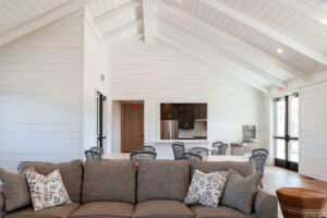 A modern, open-concept living space in Lakeview Village features a gray sofa in the foreground, a dining table with six chairs, a kitchen area, and white shiplap walls with a vaulted ceiling.