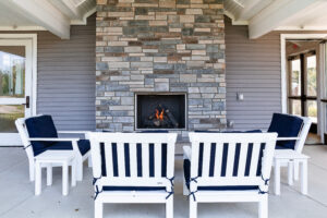 Four white wooden chairs with navy cushions are arranged around a lit stone fireplace on a covered patio with gray siding, offering cozy outdoor relaxation in Lakeview Village.