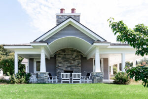A modern house in Lakeview Village with gray siding, a large stone chimney, white trim, and a covered porch with white chairs on a green lawn.