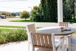 A round patio table with four chairs sits on a porch at Lakeview Village, overlooking a road, trees, and a lake in the background. A mug and newspaper rest on the table.