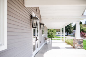 A covered front porch with white pillars, gray siding, large lantern-style lights, and a bench offers charming Lakeview Village vibes with a peaceful view of the yard and street.