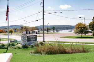 A Lakeview Village sign by Aspire stands near a road, framed by a sparkling lake, trees, hills, and boats in the background on a sunny day.