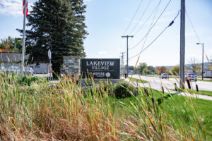 A sign reading "Lakeview Village, Aspire" stands by a roadside in Lakeview Village, with grass, power lines, trees, and passing vehicles visible in the background.