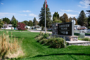 A sign reading "Lakeview Village by Aspire" stands on a stone base, welcoming you to Lakeview Village—a community of mobile homes with green lawns and shady trees.