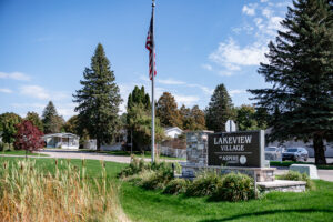 A stone and metal sign reading “Lakeview Village by Aspire” stands near an American flag, green lawn, trees, and residential homes beneath a clear blue sky in Lakeview Village.
