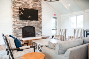 A modern Lakeview Village living room with a stone fireplace, wall-mounted TV, striped armchairs, a gray sofa, and a wooden coffee table. Natural light enters through windows and a glass door.