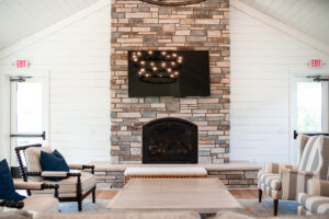 A cozy Lakeview Village living room features a stone fireplace with a wall-mounted TV above, two armchairs, a sofa, and a wooden coffee table.
