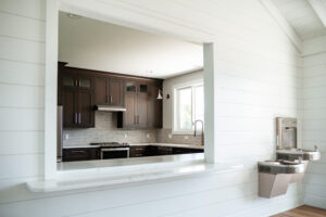 Modern kitchen with dark cabinets, stainless steel appliances, and a marble counter, viewed through a large open interior window in Lakeview Village; water fountain graces the adjacent white wall.