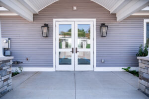Double glass doors with white framing and black handles, flanked by two large lantern lights, create a welcoming entrance to this Lakeview Village home with gray horizontal siding and elegant stone columns.