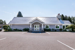 Single-story building with gray siding, a metal roof, and a central entrance, set in front of an empty parking lot at Lakeview Village on a clear day.