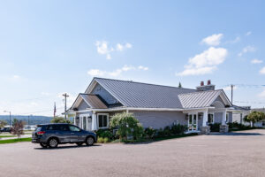 A single-story building with a metal roof and stone accents stands in Lakeview Village, a dark SUV parked in front beneath the clear sky.