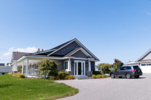 A single-story gray house with a covered porch, white trim, and a dark roof sits in Lakeview Village, next to a parked SUV in the driveway on a sunny day.