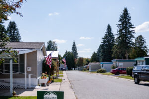A quiet street in Lakeview Village mobile home park, lined with manufactured homes, several American flags, parked vehicles, and tall trees under a clear sky.