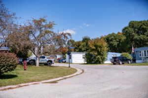 A curved street in Lakeview Village mobile home park with several parked vehicles, trees, and white manufactured homes on a sunny day.