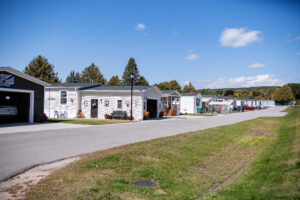 A row of manufactured homes lines a paved street in Lakeview Village, a residential community with lawns and trees under a clear blue sky.