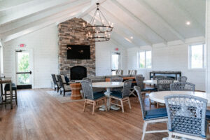 A bright, modern lounge at Lakeview Village with white walls, wooden floors, a stone fireplace, a TV, round tables with wicker chairs, and large windows letting in natural light.