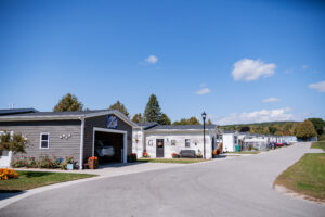 A row of manufactured homes with driveways at Lakeview Village on a clear, sunny day; a car is parked in one garage, and trees are visible in the background.