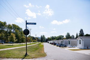 A street in Lakeview Village with manufactured homes on one side and a Meadow Ln street sign in the foreground, all under a clear blue sky.