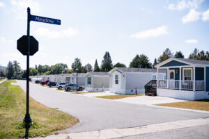Row of manufactured homes line Meadow Lane in Lakeview Village, with parked cars and trees in the background beneath a partly cloudy sky.