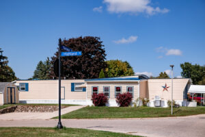 A beige manufactured home with red-trimmed windows sits at the corner of Teresa and Highland Drive in Lakeview Village on a sunny day, featuring a star decoration on the wall and shrubs in front.