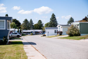 A quiet residential street in Lakeview Village features mobile homes on both sides, green lawns, parked cars, and trees under a blue sky with scattered clouds.