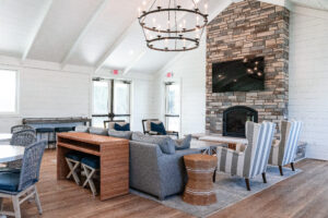 A modern Lakeview Village living room featuring a stone fireplace, mounted TV, gray sectional sofa, striped armchairs, and wood flooring beneath a large chandelier.