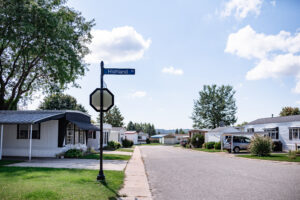 A quiet street in Lakeview Village mobile home park with a "Highland" street sign, lined with trees and manufactured homes under a partly cloudy sky.