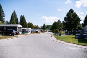 Wide paved street in Lakeview Village lined with mobile homes, parked cars, green lawns, and tall pine trees under a partly cloudy blue sky.