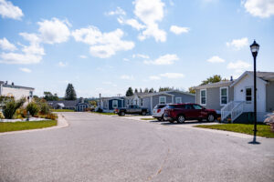 A suburban street in Lakeview Village is lined with manufactured homes, parked cars, and a streetlamp under a bright, partly cloudy sky.