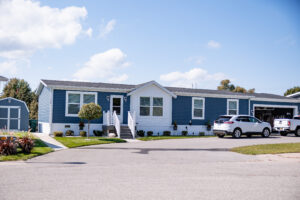 Single-story blue and white manufactured home located in Lakeview Village, featuring front steps, parked cars in the driveway, and small trees on a sunny day.