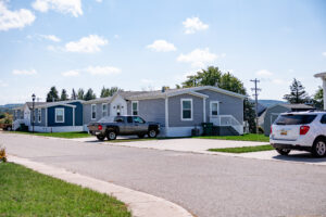 A suburban street in Lakeview Village with manufactured homes, a parked pickup truck and SUV, green lawns, and clear skies.