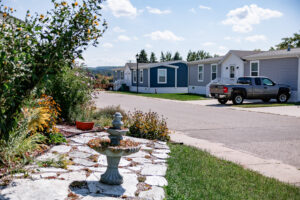 A row of mobile homes lines a paved street in Lakeview Village, with a parked pickup truck and a landscaped garden featuring a small birdbath in the foreground.