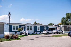 Row of manufactured homes with driveways in Lakeview Village, two parked cars, a lawn, and lamppost on a sunny day.