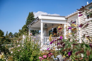 A beige house with a covered front porch and white railing sits in Lakeview Village, surrounded by a garden of blooming flowers under a clear blue sky.